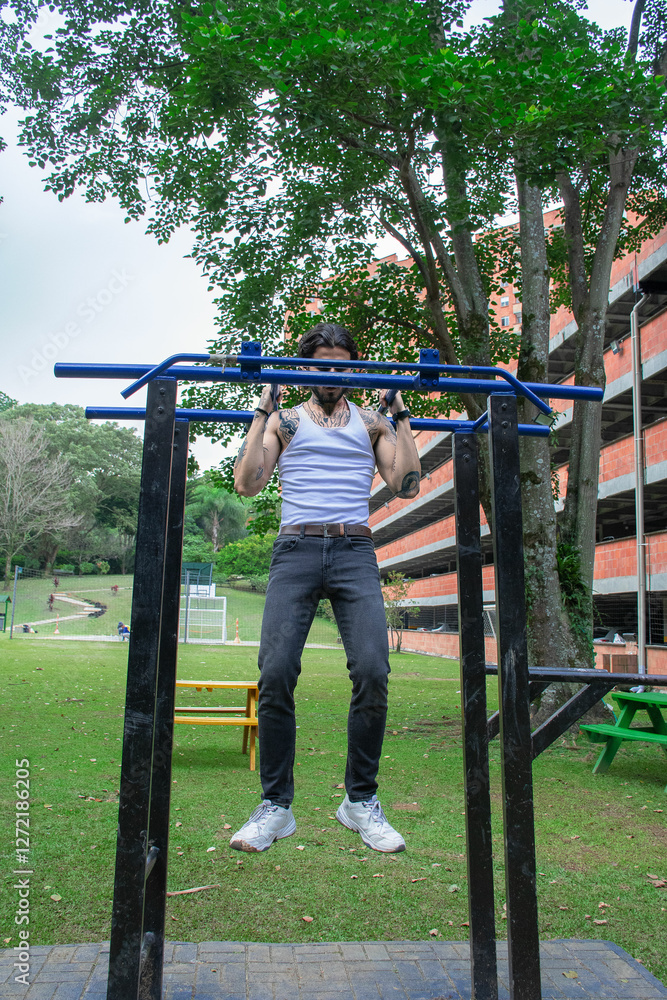 Obraz premium Young tattooed man doing pull-ups and parallel bars exercise in a public park surrounded by nature