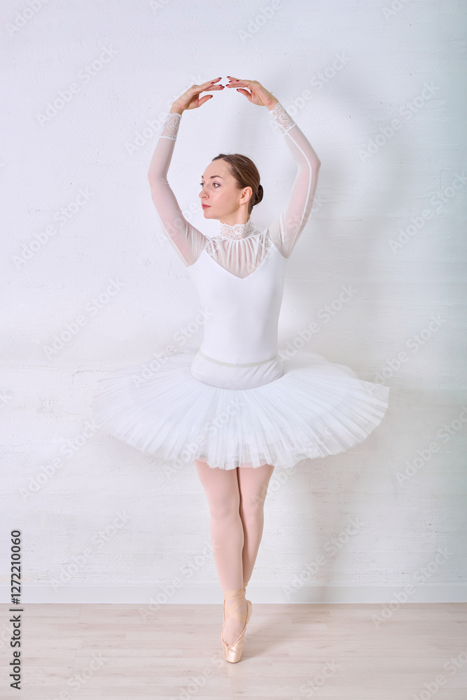 Naklejka premium Young female ballerina in elegant white tutu stands gracefully on pointe in bright minimalist studio. Soft lighting highlights serene expression and delicate posture against light-toned background