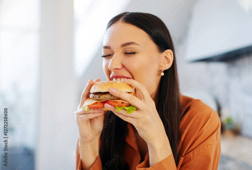 Satisfied hungry lady biting tasty burger with pleasure, enjoying cheat meal for lunch in kitchen interior, closeup Satisfied hungry lady biting tasty burger with pleasure, enjoying cheat meal for lunch in kitchen interior, closeup
