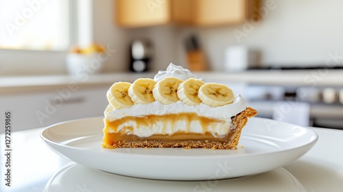 Banoffee pie slice showing layers of biscuit, caramel, cream and banana on white plate in kitchen