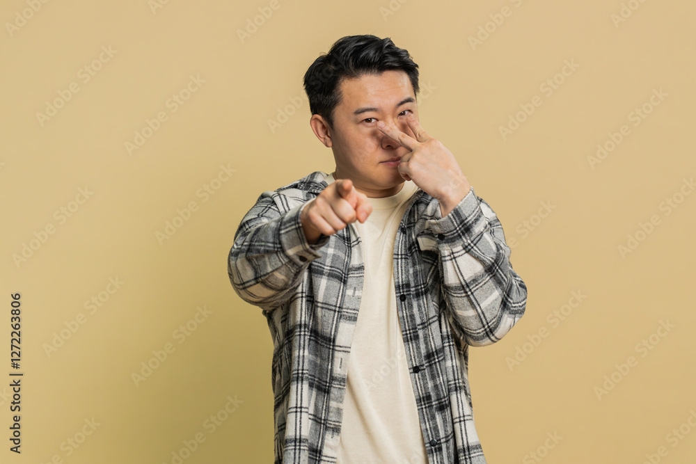 Fototapeta premium I am watching you. Confident attentive Asian middle-aged man pointing at eyes and camera, show I am watching you gesture, spying on someone. Strict adult Chinese guy isolated on beige background