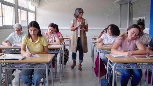 High school students are taking a test in a classroom while the teacher watches them and walks around the room talking to them. Video with copy space.