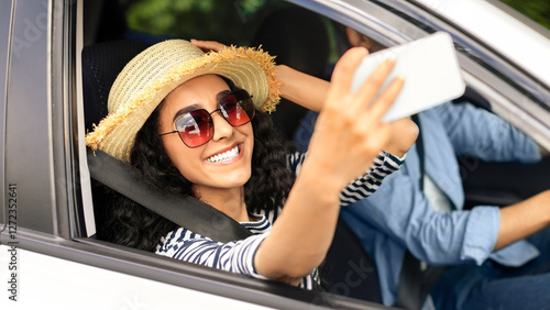 Positive young woman taking selfie on mobile phone while having car trip with boyfriend, sharing happy photos with friends on social media, wearing summer hat and sunglasses, travelling concept