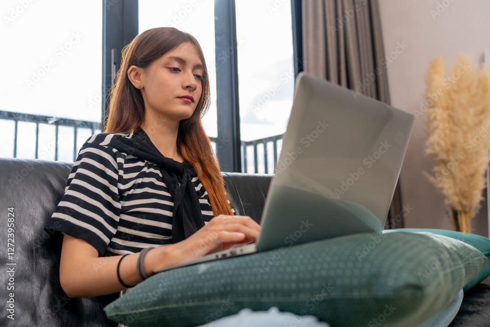 Naklejka premium young woman works on laptop while sitting on couch, focused and engaged. modern interior features large windows and decorative plants, creating cozy atmosphere