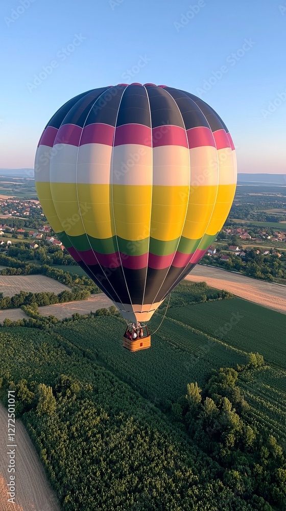 Obraz premium Colorful hot air balloon soaring over green fields and distant village at sunset