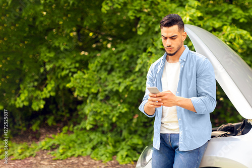 Puzzled middle-eastern guy driver standing by broken car, using cellphone, watching videos on Internet, trying to fix automobile, young arab man looking for auto service, copy space photo