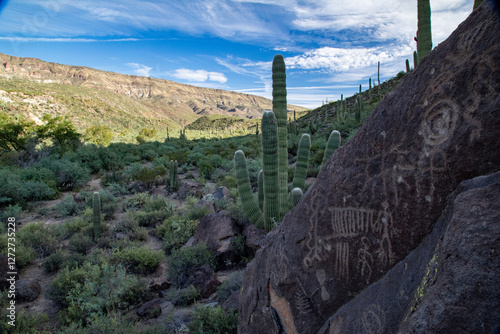 Ancient petroglyphs etched into a desert boulder stand against a stunning Sonoran landscape, featuring towering saguaro cacti, rugged mountains, and a vast blue sky filled with wispy clouds.