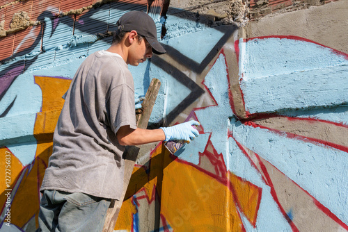 Young graffiti artist painting a colorful mural on a concrete wall using spray paint and wearing protective gloves