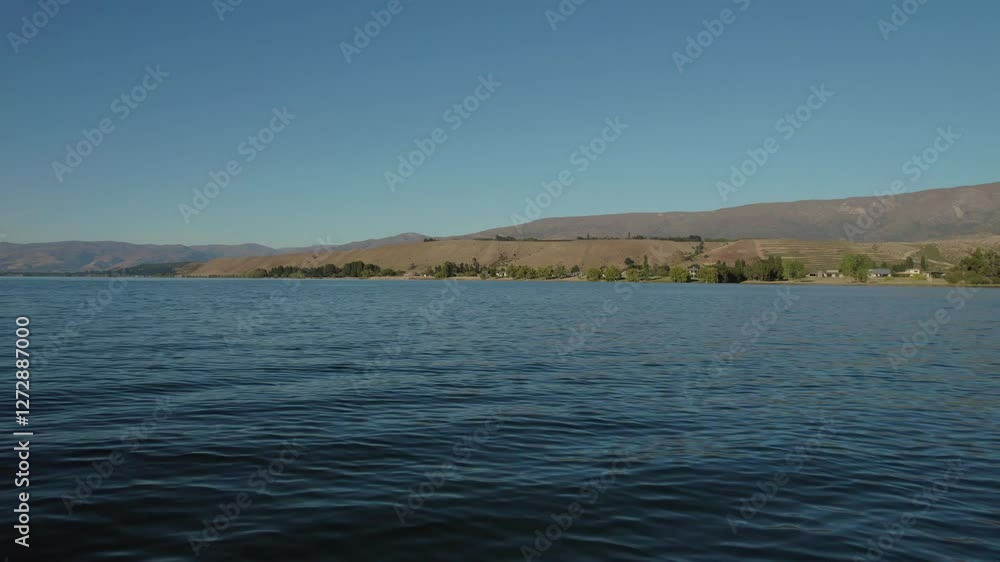 Low-altitude aerial view speeding along the water's edge near Cromwell, New Zealand. The tranquil waters, surrounding hills, and green landscape create a picturesque, peaceful scene. Low-altitude aerial view speeding along the water's edge near Cromwell, New Zealand. The tranquil waters, surrounding hills, and green landscape create a picturesque, peaceful scene.