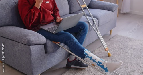 Unknown man with leg injury working distantly, types on laptop and answers call on smartphone, poses on sofa with crutches nearby, recovering at home. Rehabilitation andd health concept