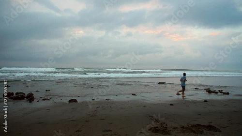 A young man jogs along the sandy beach at sunrise for exercise, captured at Echo Beach in Bali. The scene features soft morning light, gentle waves, and a peaceful coastal setting