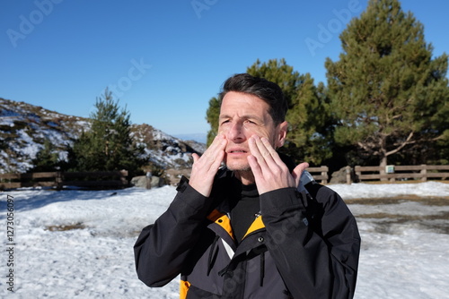 A man outdoors in a snowy landscape, wearing a black winter jacket, applying sunscreen to his face with closed eyes, protecting his skin from the sun at high altitude.