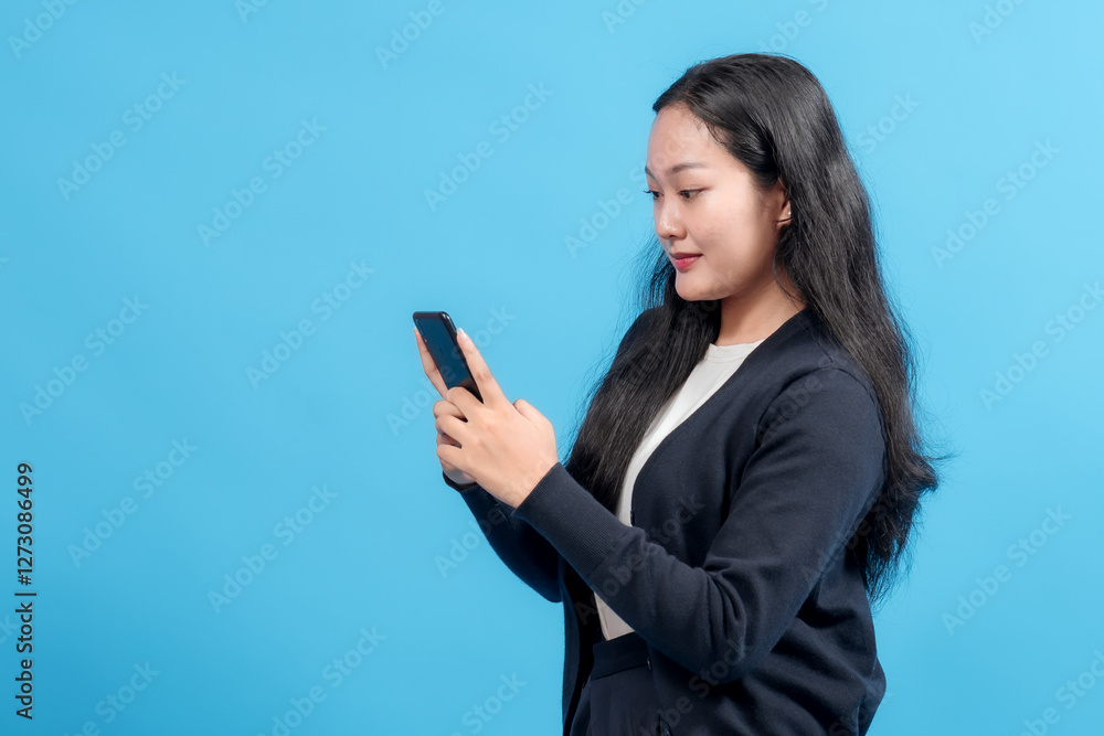 A focused woman wearing a dark sweater stands and interacts with a smartphone against a bright blue studio backdrop. A focused woman wearing a dark sweater stands and interacts with a smartphone against a bright blue studio backdrop.