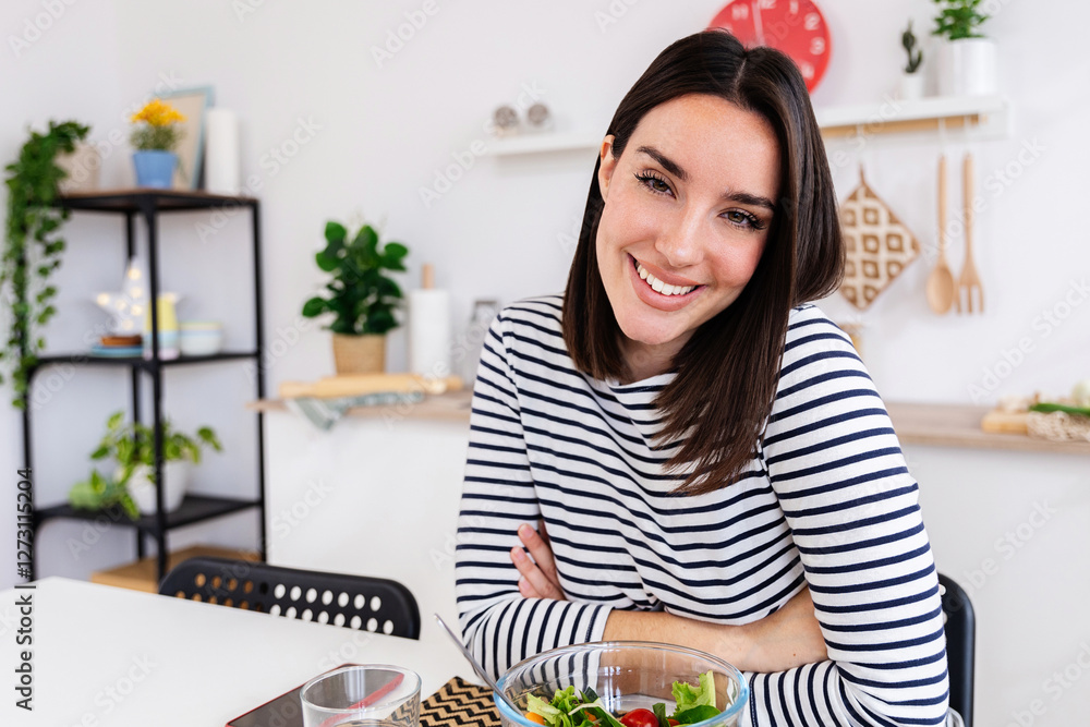 Young happy beautiful woman in a striped shirt smiling at camera with a bowl of salad sitting in the kitchen at home Young happy beautiful woman in a striped shirt smiling at camera with a bowl of salad sitting in the kitchen at home