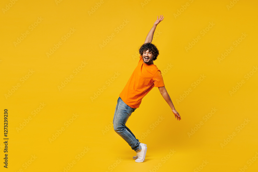 Full body young cheerful Indian man wear orange t-shirt casual clothes standing on toes dancing lean back have fun spread hands looking camera isolated on plain yellow background. Lifestyle concept. Full body young cheerful Indian man wear orange t-shirt casual clothes standing on toes dancing lean back have fun spread hands looking camera isolated on plain yellow background. Lifestyle concept.