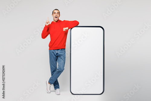 Full body young man he wear red hoody casual clothes big huge blank screen mobile cell phone smartphone with workspace copy space area point index finger up isolated on plain solid white background. photo