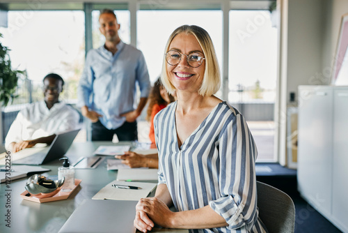 Smiling businesswoman Leading Diverse Meeting in Modern Office. She leans over the table as she looks into the camera and smiles. Her three diverse colleagues look smilingly in the same direction