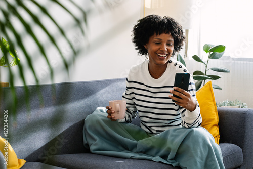Wallpaper Mural Young happy woman using mobile phone while enjoying hot tea or coffee drink relaxing on the sofa in a cozy winter day at home Torontodigital.ca