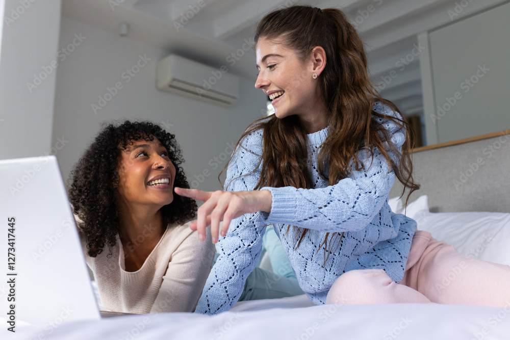 Two women relaxing on bed, one pointing at laptop screen while other smiling Two women relaxing on bed, one pointing at laptop screen while other smiling