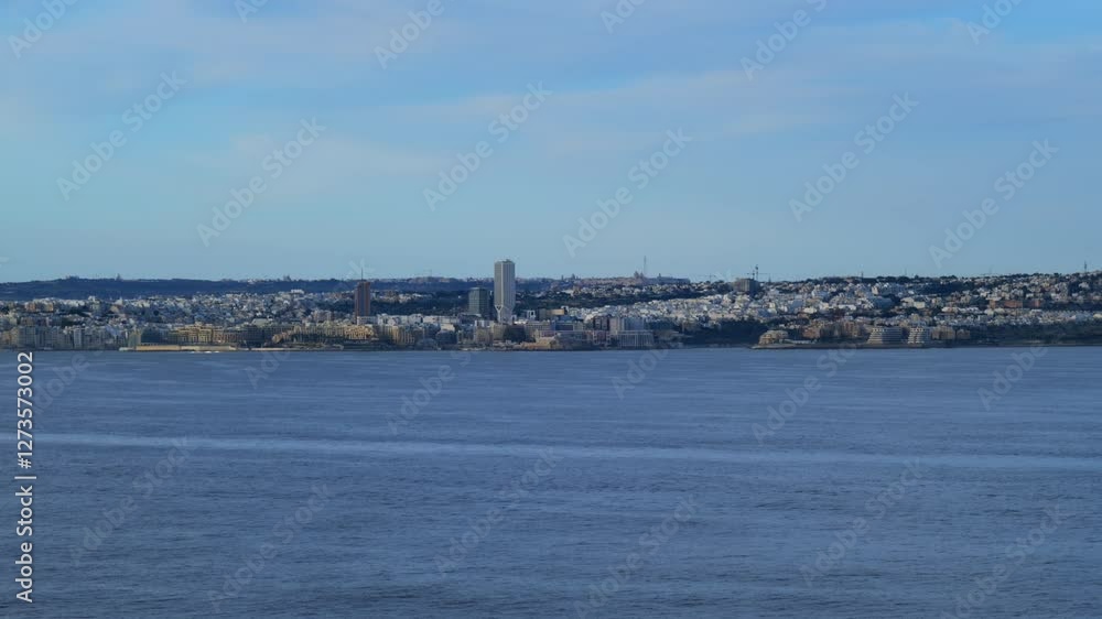 The skyline of St. Julian’s, Malta, with Mercury Tower and modern buildings is visible from the sea. The deep blue water stretches towards the coast, reflecting the clear sky in the background.