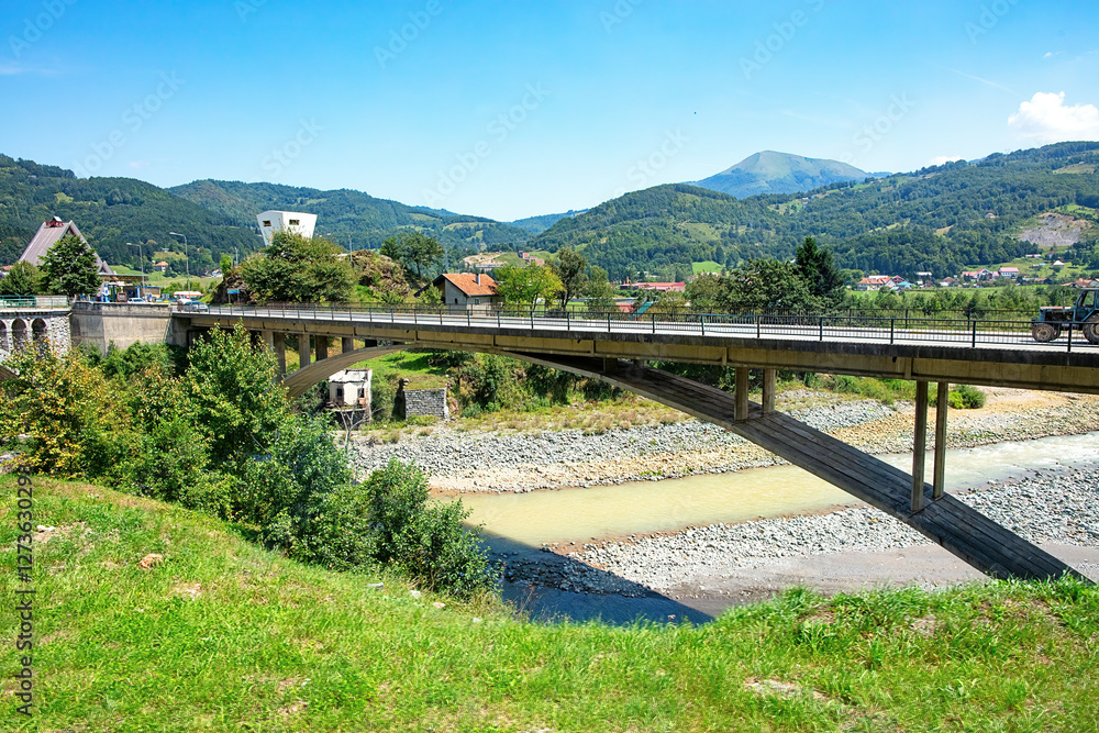 Scenic bridge crossing a serene river with lush mountains in the background on a sunny day