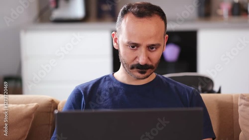 A focused young Turkish man uses his black laptop in his living room wearing a navy blue t-shirt photo