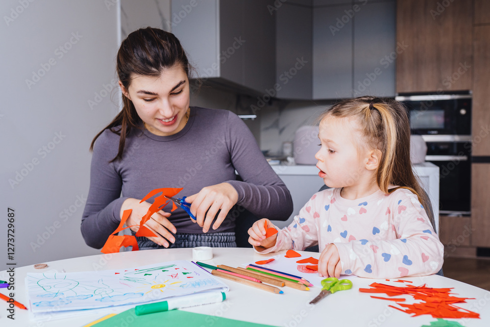 mother and 4-year-old daughter sit at white table in cozy modern kitchen, making crafts together. Child in heart-patterned pajamas focused on cutting paper hearts, mom gently helps creating. mother and 4-year-old daughter sit at white table in cozy modern kitchen, making crafts together. Child in heart-patterned pajamas focused on cutting paper hearts, mom gently helps creating.