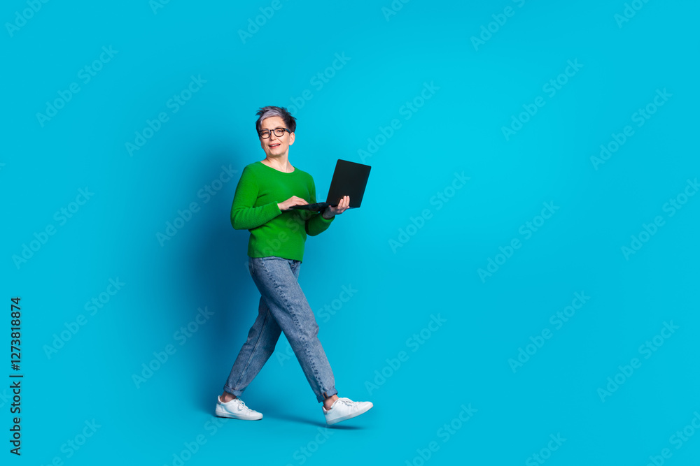 Joyful senior woman with laptop stands against blue background showcasing stylish green sweater and jeans Joyful senior woman with laptop stands against blue background showcasing stylish green sweater and jeans