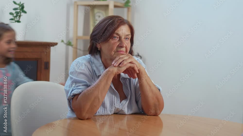Thoughtful senior woman sitting at wooden table while granddaughter playfully covers her eyes from behind, embracing and kissing her, spending leisure time together at home