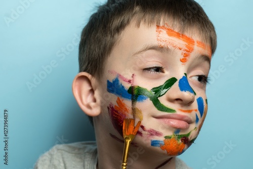 Child with colorful face paint holding a paintbrush, blue background. Perfect for themes of creativity, childhood art, playfulness, and artistic exploration
