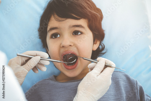 Cute arab boy sitting at dental chair with open mouth during oral checking up with doctor. Visiting sunny dentist office