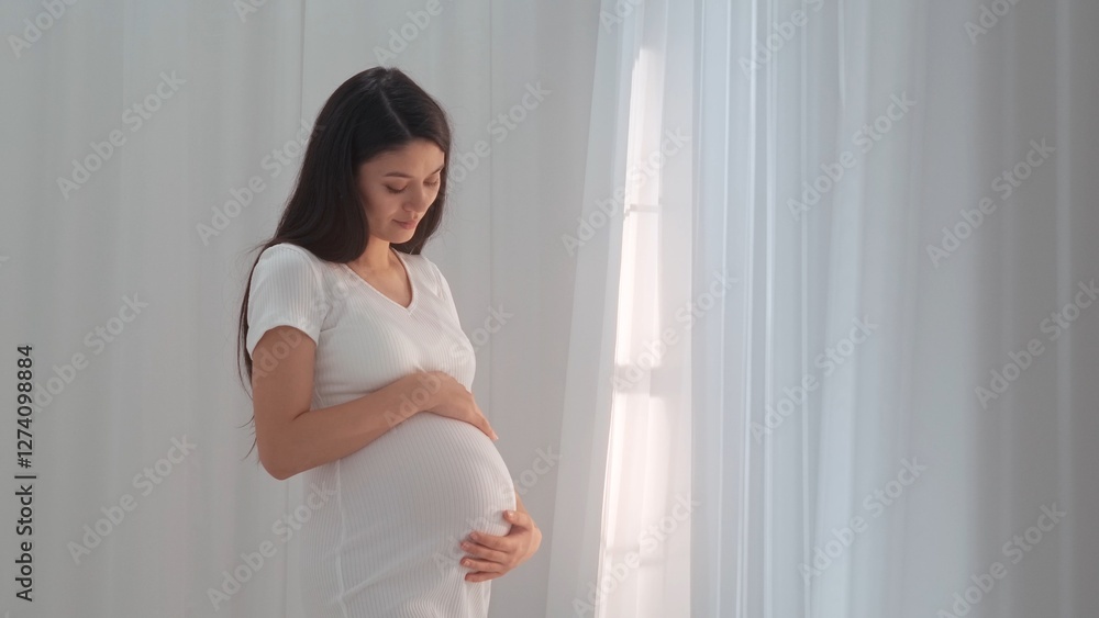 Young happy Asian pregnant woman posing in studio with white curtains in background Young happy Asian pregnant woman posing in studio with white curtains in background