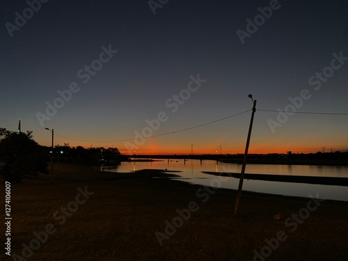 A calm river at sunset, reflecting the orange sky, with a leaning pole and bridge in the distance
