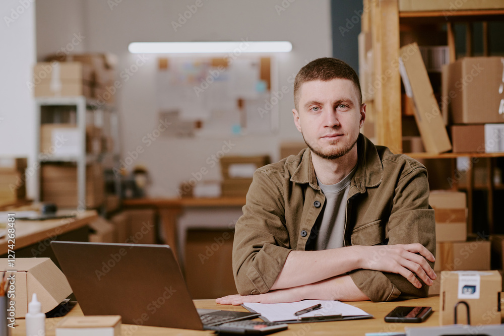 Portrait of man sitting at desk. Engaging in various office tasks with arms crossed surrounded by boxes and office materials creating a busy atmosphere Portrait of man sitting at desk. Engaging in various office tasks with arms crossed surrounded by boxes and office materials creating a busy atmosphere