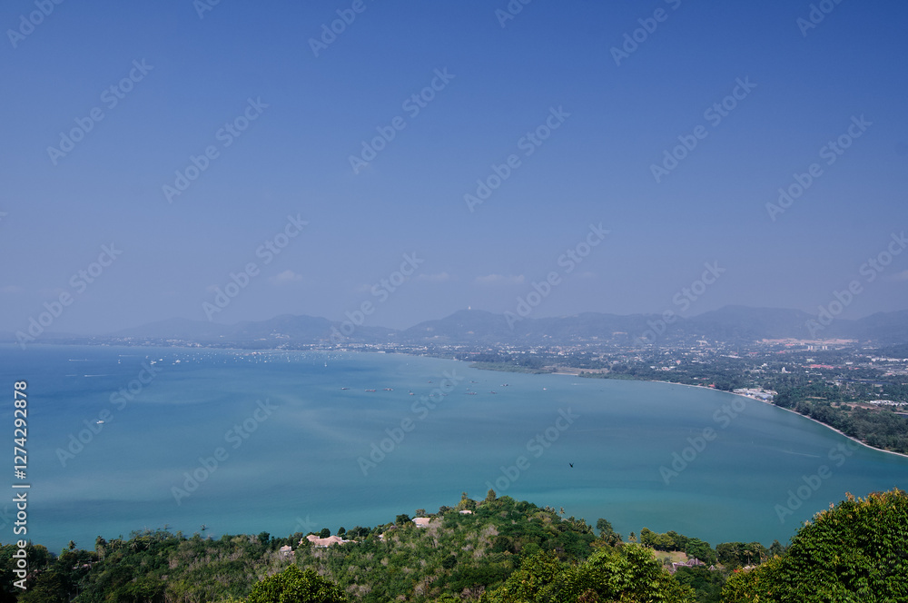A scenic view of a coastal Thailand landscape with a calm blue sea, lush green hills, and a clear blue sky. The horizon shows distant mountains and a few scattered clouds. A scenic view of a coastal Thailand landscape with a calm blue sea, lush green hills, and a clear blue sky. The horizon shows distant mountains and a few scattered clouds.