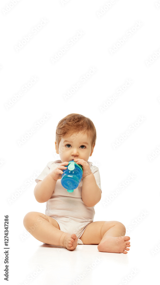 Little baby, dressed white onesie, sitting and drinking from blue sippy cup while holding it with both hands against white background. Concept of childhood, nourishment, baby care, healthy food. Little baby, dressed white onesie, sitting and drinking from blue sippy cup while holding it with both hands against white background. Concept of childhood, nourishment, baby care, healthy food.