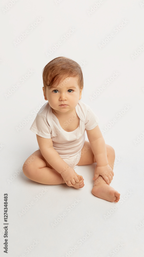 Curious little baby wearing white onesie sitting on floor holding tiny foot while gazing forward against white studio background. Concept of childhood, growth, exploration, natural curiosity. Curious little baby wearing white onesie sitting on floor holding tiny foot while gazing forward against white studio background. Concept of childhood, growth, exploration, natural curiosity.