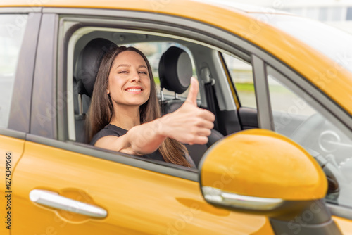 Obraz na płótnie A young woman with long brown hair and a bright smile sits in the driver's seat of a yellow car, giving a thumbs-up gesture through the open window with a confident and positive expression. w salonie