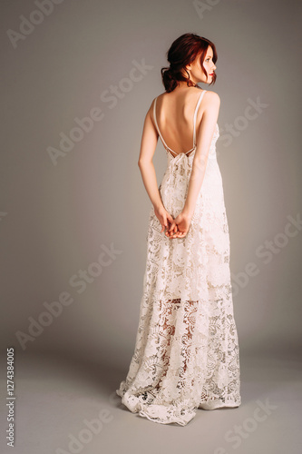 Young redhead woman's studio portrait. Waisted transparent maxi dress. White backless evening outfit. Sleeveless wedding gown with straps and long skirt. Female summer fashion, occasion look,rear view