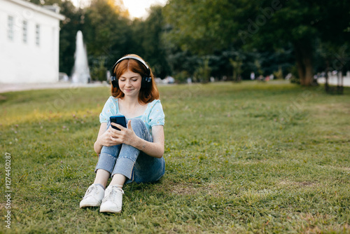 Teenage girl listening music through wireless headphones and smartphone sitting on the grass