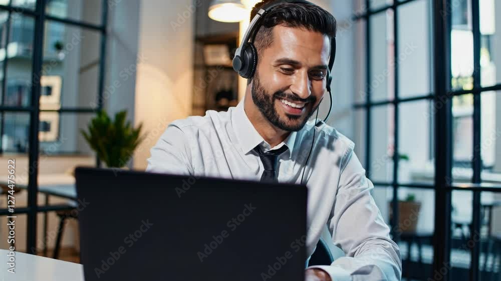Professional video call setting with a man in a headset, smiling at a laptop. Shot from a low angle in a modern office environment. Professional video call setting with a man in a headset, smiling at a laptop. Shot from a low angle in a modern office environment.