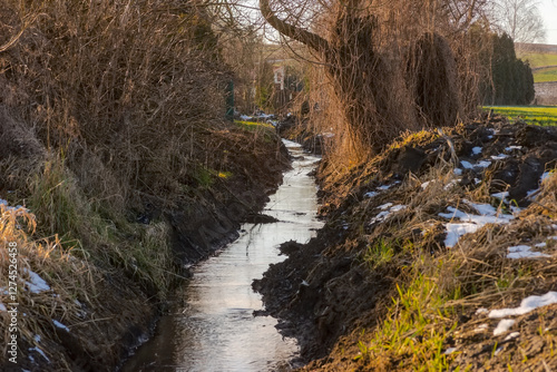 Strumień płynący przez pola w słoneczny zimowy dzień. Wąski strumień wije się przez brązowe, błotniste pola, otoczone rzadką zimową roślinnością. Na ziemi rozrzucone są łaty śniegu, co sugeruje opad  photo