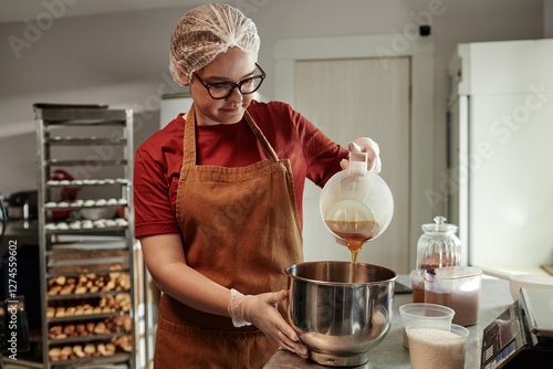 Medium shot of young female baker wearing hairnet pouring maple syrup into metal bowl mixing ingredients while baking artisan bread in bakehouse kitchen