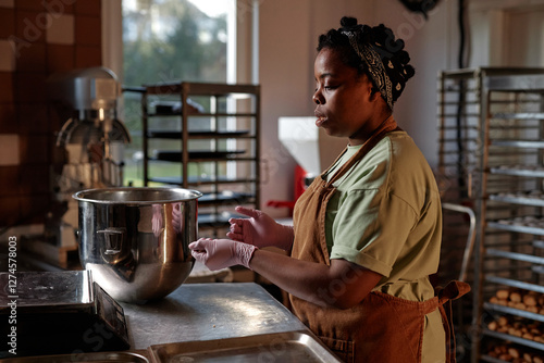 Side view of focused Black female baker pre shaping raw dough to make cookies at working station in small bakery kitchen photo