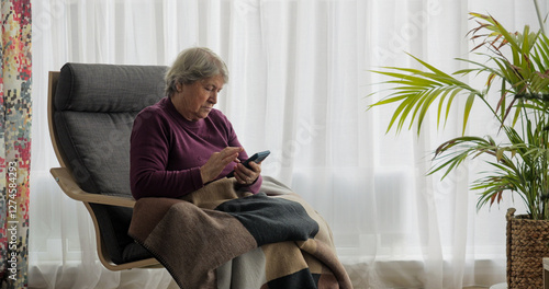 Senior woman using mobile phone sitting on the armchair in the living room