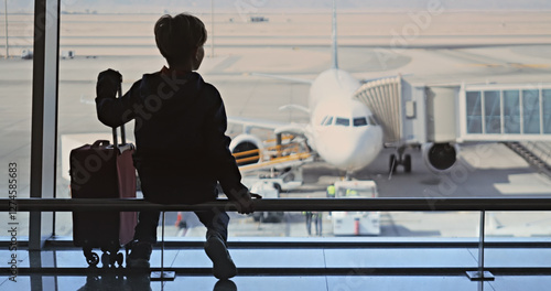 Little boy with suitcase watching airplane from large window of airport