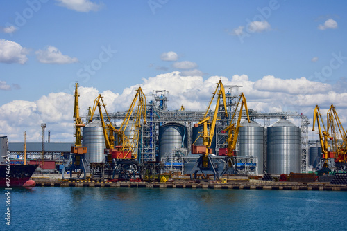 Cranes and silos are actively being constructed at a busy shipping port as ships await their cargo under a sunny sky