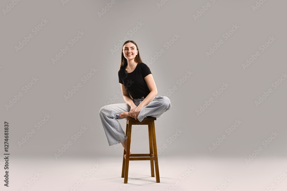 Smiling woman in stylish jeans sitting on stool against grey background Smiling woman in stylish jeans sitting on stool against grey background