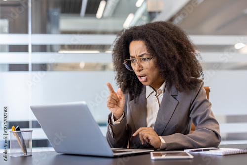 Angry young african american business woman sitting at desk in office and talking angrily on video call on laptop camera while pointing finger at screen