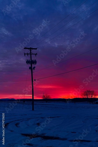 An electrical pole stands against a sky with red and blue tones. The snowy ground reflects the evening light. Rural landscape with trees